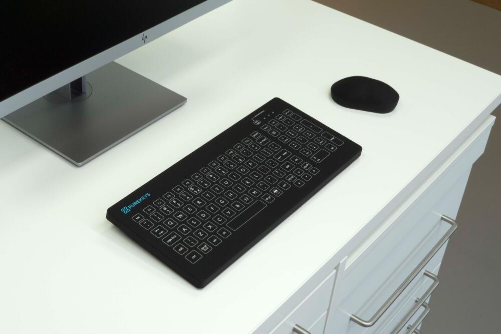Purekeys medisch toetsenbord US zwart op medische werkplek naast monitor en muis; vlak, hygiënisch en eenvoudig te reinigen.