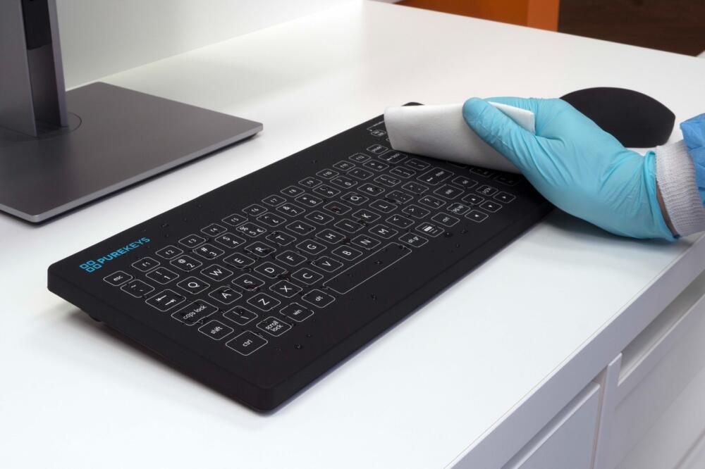 Hand met handschoen maakt Purekeys medisch toetsenbord US zwart schoon met doek; vlak oppervlak geschikt voor desinfectie.