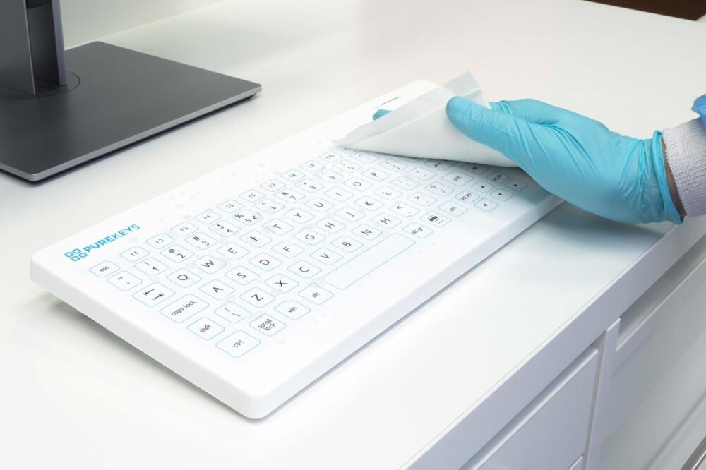 Hand met handschoen reinigt het Purekeys medisch toetsenbord wit met een doek; vlak oppervlak geschikt voor desinfectie.